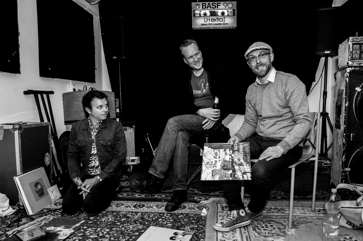 Drie mannen ontspannen in muziekstudio met instrumenten.