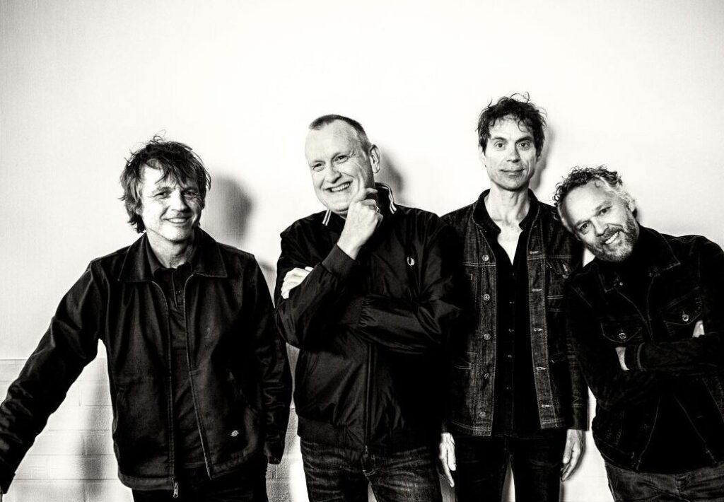 Vier mannen poseren tegen witte muur in zwart-wit.