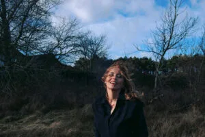 Vrouw lacht in winderig landschap onder bewolkte hemel.