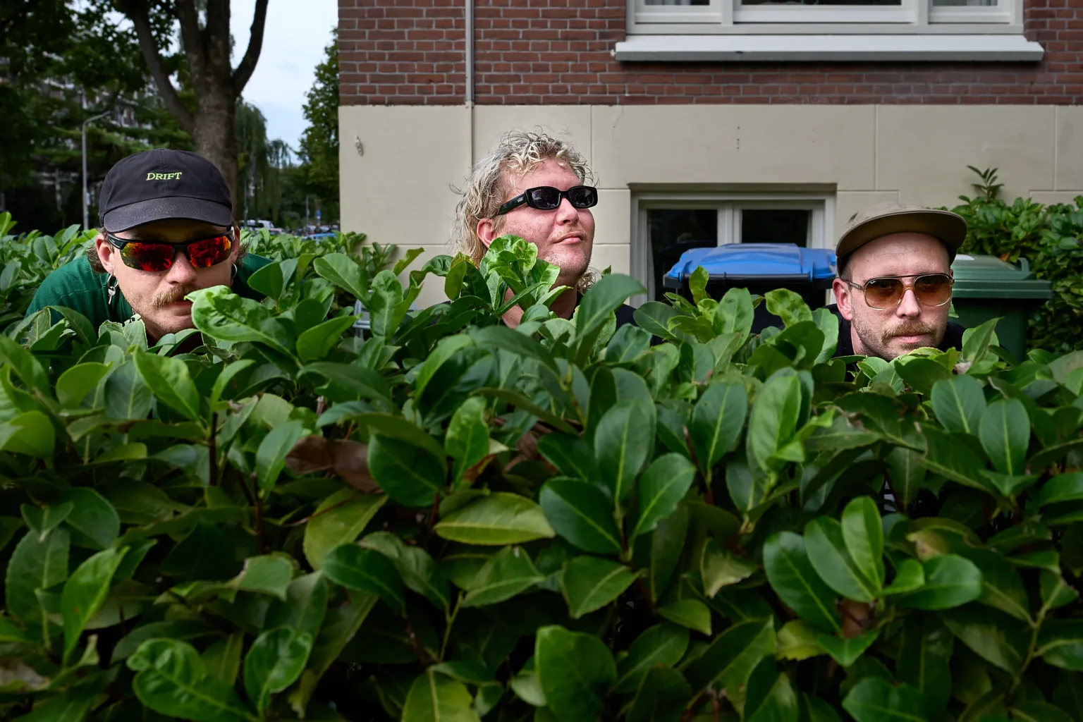 Drie mannen kijken over hoge heg bij huis.