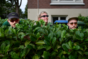 Drie mannen kijken over hoge heg bij huis.